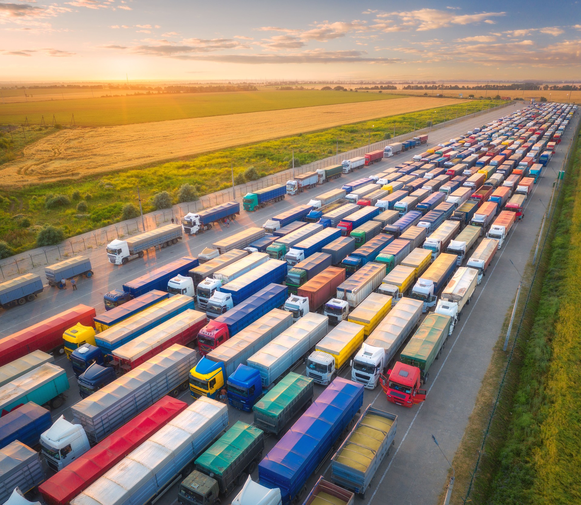 Aerial view of colorful trucks in terminal at sunset in summer. Top view of logistic center. Transportation. Cargo transport, shipping. International trucking. View from drone of trucks, road, field